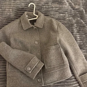 Grå ulljacka med stora fickor - Stilren grå jacka i ullmix med raka linjer och stora framfickor. Kortare modell med klassisk krage och knäppning framtill. Perfekt för lager på lager och enkel att matcha till olika outfits.