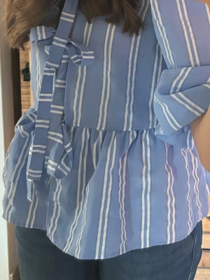Blåvit randig blus med knytband - Snygg blå blus med vita vertikala ränder och volang nedtill. Blusen har långa ärmar och ett dekorativt knytband vid halsen. Perfekt till jeans för en fräsch look. Materialet känns lätt och luftigt. Perfekt till sommaren köpt från only köpt för 500