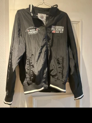 Svart vindjacka från Saint Vincent - Snygg svart vindjacka från Saint Vincent med vita detaljer och dragkedja. Jackan har broderade loggor på bröstet, ribbade muddar och krage samt två fickor fram. Materialet är glansigt och lätt, perfekt för blåsiga dagar.