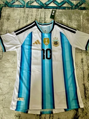 Argentina Messi matchtröja Adidas - Säljer en officiell Argentina matchtröja från Adidas med Messi och nummer 10 på ryggen. Tröjan är vit med blå ränder, har svarta detaljer på axlarna och guldiga loggor framtill.   ^Tröjan är player version^