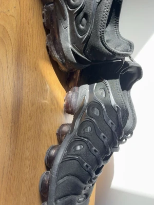 Nike Air Vapormax Plus svarta sneakers - Svarta Nike Air Vapormax Plus sneakers med ikonisk bubblig sula och detaljer i plast längs ovandelen. Skorna har en sportig siluett, meshmaterial och diskret Swoosh-logga på sidan. Perfekta för dig som gillar streetwear och vill ha något som sticker ut.
