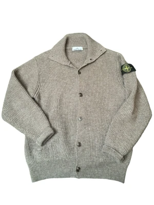 Beige kofta från Stone Island - Riktigt snygg beige kofta från Stone Island med ribbstickad struktur och klassisk logotyp på ärmen. Koftan har knappar framtill, bred krage och långa ärmar. Perfekt för dig som gillar stilrena och exklusiva plagg. Tröjan är L men passar som en M (tyvärr inga tags i tröjan) men den känns som kvalitet sömmarna ser bra ut och även badgen ser ut å vara äkta, Tröjan har inga fläckar eller hål men är lite smått norpig