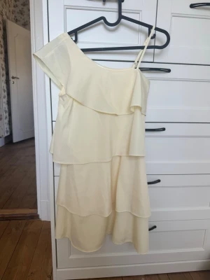 Ljusgul oneshoulder volangklänning - Söt ljusgul kortklänning med oneshoulder och volangdetaljer. Klänningen har ett axelband och flera lager volanger som ger en flowy känsla. Perfekt för sommarens alla tillfällen och ger en fräsch look. I storlek 170 men passar en XS/S