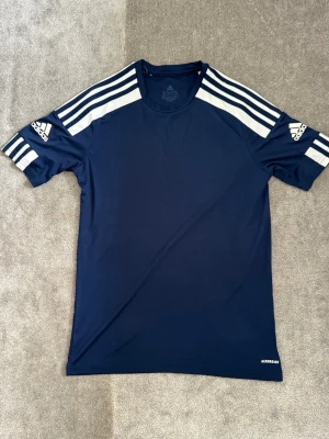 Mörkblå Adidas träningströja - Snygg mörkblå träningströja från Adidas med vita ränder på axlarna och logga på båda ärmarna. Tillverkad i lätt och snabbtorkande material (Aeroready), perfekt för träning eller sportaktiviteter. Klassisk rund hals och korta ärmar.