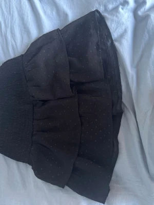 Svart volangkjol med prickigt mönster - Supersöt svart kortkjol med volanger och diskret prickigt mönster. Kjolen har en bred, smockad midja som ger en bekväm och snygg passform. Perfekt att styla med en enkel topp eller hoodie för en trendig look.