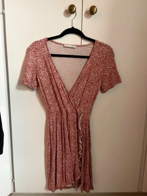 Röd omlottklänning med blommönster - Söt röd omlottklänning med små vita blommor och korta ärmar. Klänningen har v-ringning och är tillverkad i ett lätt, mjukt tyg som faller fint. Perfekt för dig som gillar en feminin och avslappnad stil.