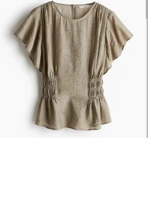 Beige blus med volang och rynk - Snygg grön blus med korta, vida ärmar och volang nertill. Blusen har rynkade detaljer i midjan som ger en smickrande passform. Perfekt för en trendig look.