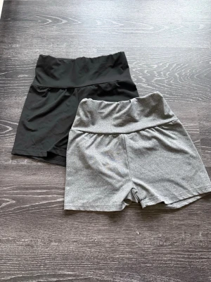 Två par träningshorts - Två par högmidjade shorts ( går att vika ner) , ett par i svart och ett par i ljusgrått. Båda har bred linning och är tillverkade i stretchigt syntetmaterial, perfekta för träning eller avslappnade dagar.