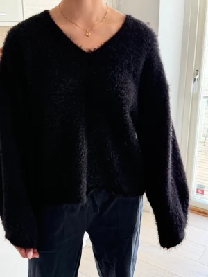 Svart fluffig stickad tröja Gina Tricot - Mysig svart stickad tröja från Gina Tricot med fluffig textur och v-ringad hals. Tröjan har långa ärmar och en avslappnad passform, perfekt för att hålla sig varm och snygg. Materialet är mjukt och gosigt🥰✨