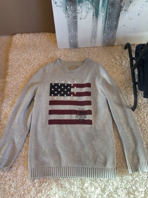 Grå stickad tröja Lexington - Säljer en grå stickad tröja från Lexington med amerikansk flagga i marinblått, vitt och vinrött på bröstet. Tröjan har rund halsringning och ribbade muddar vid ärmslut och nederkant. Perfekt för dig som gillar klassisk och avslappnad stil.
