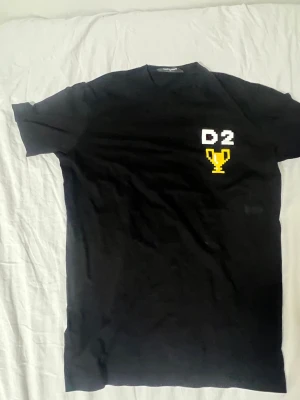 Svart Dsquared2 t-shirt med tryck - Snygg svart äkta t-shirt från Dsquared2 i storlek M. Framtill finns ett pixelinspirerat tryck med texten 'D2' och en gul pokal. Klassisk passform och rund halsringning, perfekt till jeans eller shorts.