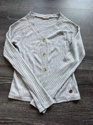 Ljusgrå ribbad kofta med knappar - Snygg ljusgrå ribbad kofta med v-ringning och träfärgade knappar framtill. Långärmad modell i mjukt material, perfekt för lager på lager. Liten rund detalj vid nederkanten. Enkel och stilren design som passar till det mesta.