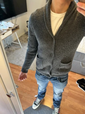 Grå kofta/Cardigan - Mysig grå kofta med sjalkrage och två stora fickor framtill. Koftan är ribbstickad och har knäppning med knappar. Perfekt att slänga på sig över en t-shirt för en avslappnad men snygg look.