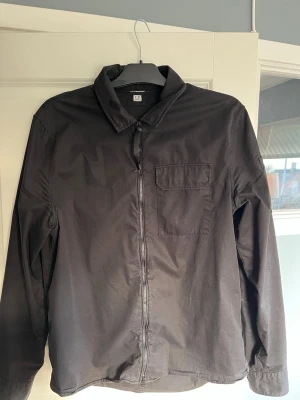 Svart overshirt från C.P. Company - Stilren svart overshirt från C.P. Company med dragkedja framtill och en bröstficka. Overshirten har klassisk krage och är tillverkad i ett lätt, slätt material som passar perfekt för lager-på-lager. Enkel och clean design som funkar till det mesta. Strl: L (men är liten i storleken). Priset är ej hugget i sten, och det är bara att komma med frågor om det finns!