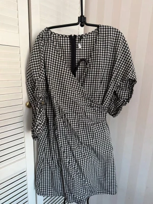 Nakd Rutig omlottklänning svartvit 36 - Kort svartvit rutig klänning med omlott-design och trekvartsärm. Klänningen har dragkedja i ryggen och puffiga ärmar för en trendig look. Perfekt för dig som gillar klassiska mönster med en modern twist.