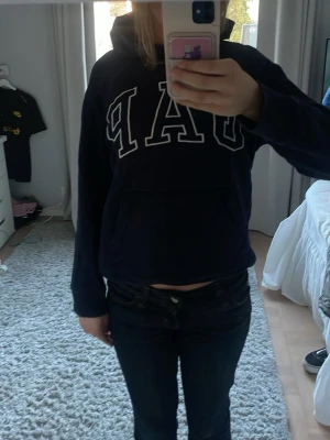 Mörkblå hoodie från GAP Kids - Mörkblå hoodie från GAP Kids med stor vit logga framtill. Tröjan har huva och en klassisk känguruficka. Perfekt för en avslappnad och sportig stil. Liten i storleken och lite korta armar.