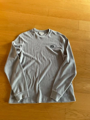 Ljusgrå ribbstickad tröja Calvin Klein - Ljusgrå långärmad ribbstickad tröja från Calvin Klein med rund halsringning och diskret logotypmärke på bröstet. Tröjan har breda muddar vid ärmsluten och är tillverkad i mjukt, stretchigt material som passar perfekt till vardagslooken. Tröjan är helt oanvänd. Storlek S men är stor i storleken.