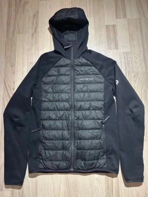 Svart hybridjacka från Peak Performance - Snygg svart cardiganjacka från Peak Performance med vadderade paneler fram och bak samt huva. Jackan har dragkedja, två sidofickor och är tillverkad i en mix av slätt tyg och quiltat material. Perfekt för lager-på-lager och streetwear.