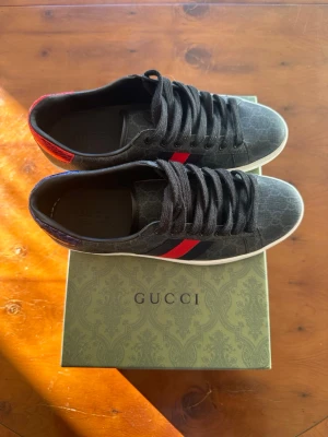 Gucci ace svarta sneakers  - Gucci sneakers i svart med monogrammönster, klassiska röda och blå ränder på sidan samt kontrasterande häl i rött och blått ormskinnsmönster. Vita sulor och svarta skosnören. Snygg och tidlös design som sticker ut.