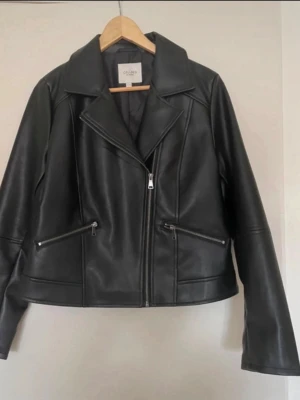 Bikerjacka  - • Bikerjacka med mjuk, följsam läderlook i polyuretan med tunt foder • Klassisk modell med silverfärgade dragkedjor i antikfinish • Ok framtill och baktill med panelsömmar runt om • Snedställda fickor framtill med dragkedjor • Långa ärmar med avskärningar och dragkedjor  St 42-44  Längd mitt bak: 60 cm  Oanvänd