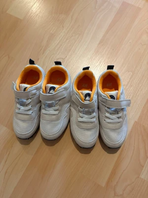 Vita sneakers från H&M med orange detalj - Snygga vita sneakers från H&M med fräscha orange detaljer på insidan. Skorna har kardborreband och elastiska snören för enkel påtagning. Ovandelen är i mesh och syntetmaterial, perfekt för en sportig och stilren look. Aldrig använda. Köp båda paren för 100kr eller ett par för 60kr!