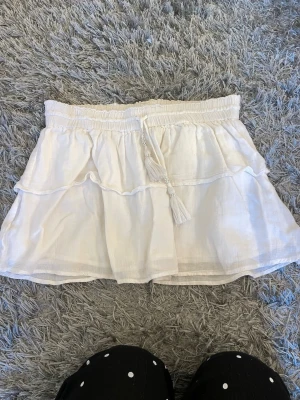 Vit volangkjol med tofsdetalj - Söt vit kjol med volanger och elastisk midja. Kjolen har en dekorativ tofs framtill och är gjord i ett lätt, luftigt tyg. Storlek 146/152 från ginas barnavdelning. Inga defekter 