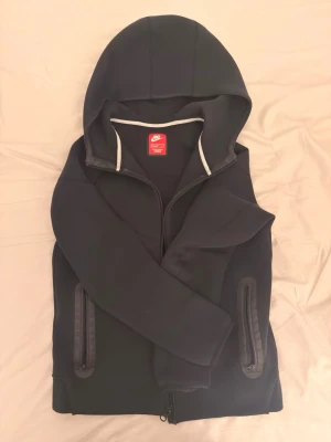 Nike tech Junior - Snygg svart hoodie från Nike med dragkedja framtill och två fickor med dragkedja. Klassisk huva och diskret vit kant på insidan av luvan. Perfekt för dig som gillar sportig och clean stil. Tillverkad i mjukt och bekvämt material.