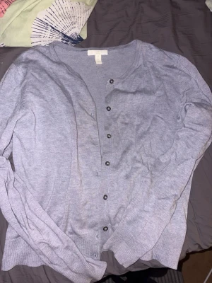 Ljusgrå-blå cardigan från H&M - Ljusgrå-blå cardigan från H&M med rund halsringning och knappar framtill. Tillverkad i ett tunt, mjukt material som passar perfekt för lager på lager. Långärmad modell med ribbade muddar vid ärmslut och nederkant.