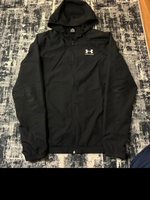 Svart hoodie från Under Armour - Säljer en svart hoodie från Under Armour med dragkedja och huva. Tröjan har en vit logga på bröstet och är tillverkad i ett lätt och sportigt material. Perfekt för dig som gillar en clean och sportig stil.