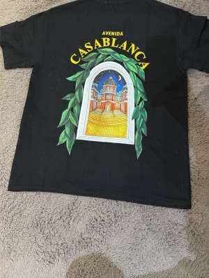 Svart Casablanca Avenida t-shirt - Svart t-shirt från Casablanca med färgglad tryckt grafik på både fram- och baksida. Motivet visar en fönsterbåge med gröna blad och en byggnad i bakgrunden, samt texten 'Avenida Casablanca' i gult. Klassisk passform och rund hals. Perfekt för dig som gillar statement-plagg.