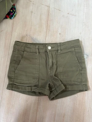 Olivgröna shorts med uppvik xxs - Snygga olivgröna shorts med uppvikta benslut och klassisk femficksdesign. Shortsen har normal passform, bälteshällor och stängs med knapp och dragkedja. Skulle passa för en xxs
