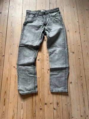 Grå raka jeans från Jack & Jones - Säljer ett par grå jeans från Jack & Jones. De är i fint skick förutom ett litet hål vid högerficka. Storleken på byxorna är 31W32L och sitter ganska löst. Hör av er vid funderingar!