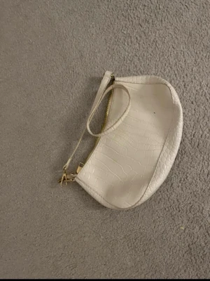 Vit axelväska i crocopräglat material - Snygg vit axelväska med crocopräglat mönster och guldfärgade detaljer. Väskan har en halvmåneform och justerbar axelrem. Perfekt för dig som gillar trendiga accessoarer och vill ha något stilrent till din outfit.