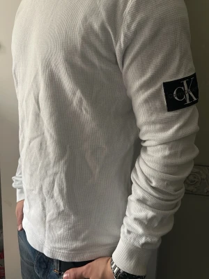 Vit långärmad tröja Calvin Klein Jeans - Säljer en stilren vit långärmad tröja från Calvin Klein Jeans. Tröjan har en diskret struktur i tyget och en svart patch med CK-logga på ärmen. Perfekt för dig som gillar enkel och clean stil.