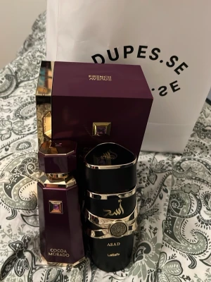 Cocoa Morado parfym helt ny och lataffa asad - Lyxig parfymflaska från French Avenue i djup lila färg med gulddetaljer och en dekorativ lila sten på både flaskan och förpackningen. Flaskan har en elegant, kantig form och ger ett exklusivt intryck. Perfekt för dig som gillar unika och snygga parfymer.