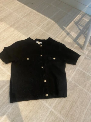 Svart kortärmad kofta Gina Tricot - Kortärmad svart kofta från Gina Tricot med dekorativa guldknappar framtill och två fejkfickor på bröstet. Koftan har rund halsringning och är stickad i mjukt material. Perfekt att styla över ett linne eller t-shirt.