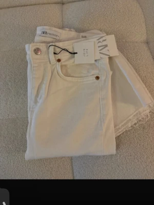 Vita raka jeans från Zara - Säljer ett par vita jeans från Zara med raka ben och rå kant nedtill. Jeansen har klassisk femficksmodell, silverfärgad knapp och detaljer i koppar. Materialet är mjukt denim i bomull. Perfekta för en clean och fräsch look.