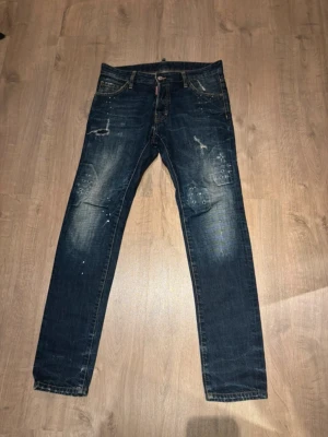 Mörkblå dsq2  - Säljer ett par mörkblå skinny jeans med slitningar och färgstänk på benen. Jeansen har klassisk femficksmodell, låg midja och smal passform. Snygga detaljer som ger en trendig och avslappnad look. Perfekta för dig som gillar streetstyle.