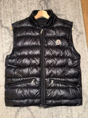Moncler Gui väst  - Snygg svart dunväst från Moncler med hög krage och två dragkedjefickor framtill. Västen har quiltad design och ett klassiskt Moncler-märke på bröstet. Perfekt att slänga över en hoodie eller tröja för extra värme. Storlek 2. Felfri