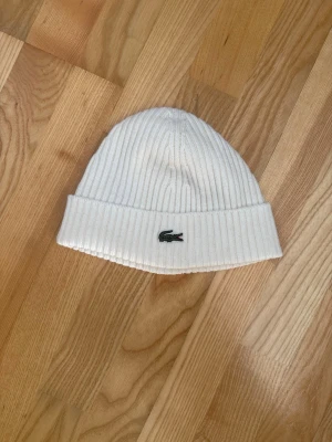 Vit ribbad mössa från Lacoste - Säljer en stilren vit ribbstickad mössa från Lacoste med den klassiska gröna krokodilloggan framtill. Mössan har uppvikt kant och är gjord i mjukt material som håller dig varm och snygg under kalla dagar.