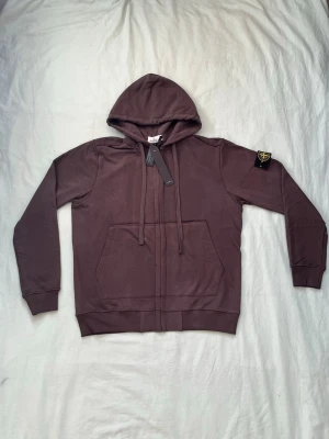 Brun hoodie från Stone Island - Snygg brun hoodie från Stone Island med dragkedja framtill, stor huva och känguruficka. Klassisk logopatch på vänster ärm och ribbade muddar vid ärmslut och nederkant. Tillverkad i mjuk bomull för skön känsla. Perfekt för en avslappnad streetwear-look.