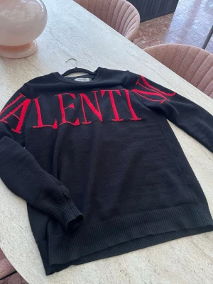 Svart stickad tröja från Valentino - Svart stickad tröja från Valentino med stora röda bokstäver över bröstet och ärmarna. Tröjan har rund halsringning och ribbstickade muddar vid ärmslut och nederkant. Perfekt för dig som gillar statement-plagg och exklusiva märken.