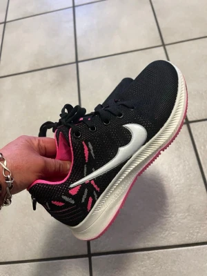 Svarta sneakers med rosa detaljer storlek 38 - Snygga svarta sneakers från PCSPORT med vita och rosa detaljer samt vit sula och rosa yttersula. Skorna har ett stickat meshmaterial, svarta skosnören och en stor vit logga på sidan. Insidan är fodrad i rosa och hälen har textdetaljer. Perfekta för en sportig look.