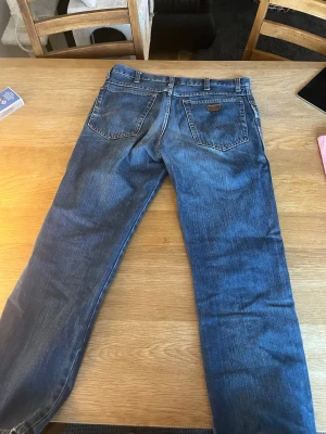 Blå raka jeans från Wrangler - Säljer ett par klassiska blå jeans från Wrangler med raka ben och femficksmodell. Jeansen har en snygg tvättad look och detaljer som Wrangler-lappen på bakfickan. Perfekta till vardags för en avslappnad stil.