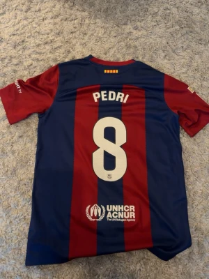 Barcelona matchtröja Pedri #8 Nike - Säljer en officiell FC Barcelona matchtröja med Pedri och nummer 8 på ryggen. Tröjan har blå och röda vertikala ränder, klubbmärke och Nike-logga på bröstet samt Spotify som sponsor. Tillverkad i lätt och ventilerande material, perfekt för fotboll eller träning.