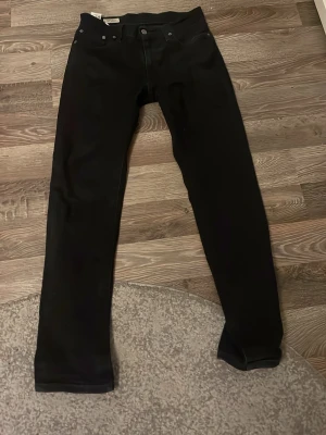 Svarta jeans från Levi's - Säljer ett par klassiska svarta jeans från Levi's med rak passform och fem fickor. Jeansen har knappgylf och är tillverkade i ett slitstarkt denimtyg. Perfekta för dig som gillar en tidlös och enkel stil.
