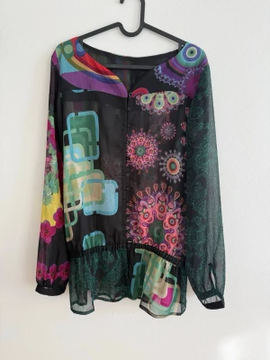 Färgstark Mönstrad Blus/Tunika  - Superfin och unik blus/tunika i tunn, luftig kvalitet med färgstarkt mönster i svart, grönt, rosa och lila. Perfekt om du gillar boho/vintage-stil ✨  Plagget har knappar framtill, långa ärmar och en lätt markerad midja med resår som ger en fin passform. Lite transparent, så passar perfekt med linne under.  Ingen storlekslapp kvar, men uppskattas till S/M beroende på hur man vill att den ska sitta.  Passar lika bra till jeans som till kjol – perfekt till både vardag och fest.