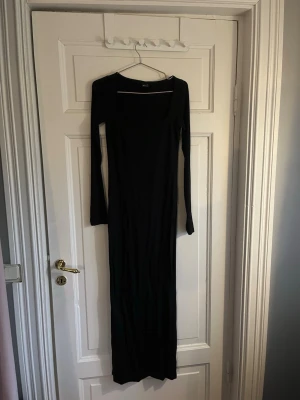 Svart långärmad långklänning - En stilren svart långklänning med lång ärm och rundad urringning. Klänningen har en figurnära passform och är tillverkad i ett mjukt, stretchigt material som ger en snygg siluett. Perfekt för dig som gillar minimalistisk och tidlös stil.