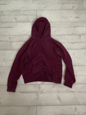 Vinröd hoodie från Gina Tricot - Säljer en vinröd hoodie från Gina Tricot med klassisk huva och ribbade muddar. Hoodien är gjord i mjukt bomullsmaterial och har en enkel, stilren design utan tryck eller dragkedja. Perfekt för en chill och avslappnad look.