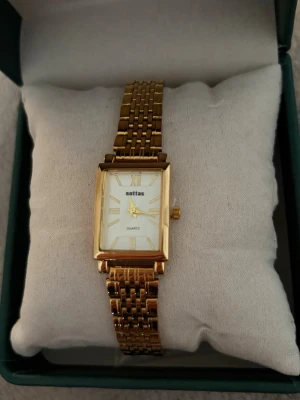 Guldfärgad rektangulär klocka Sottas - Stilren klocka från Sottas med rektangulärt ur och guldfärgad länk i metall. Urtavlan är vit med romerska siffror och tunna visare. Klassisk och elegant design som passar till många olika looks.